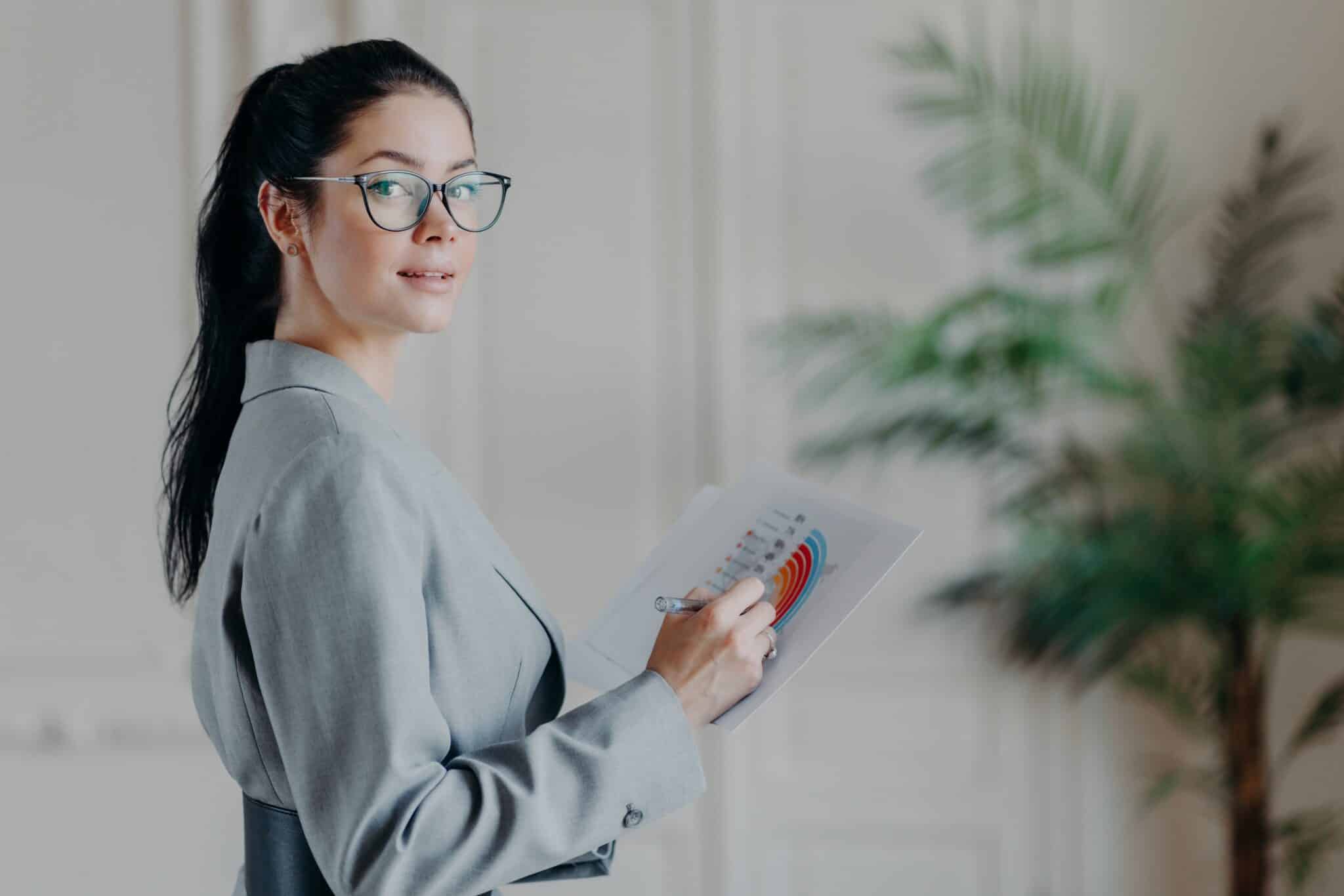 Horizontal shot of female strartuper analyzes business information, holds paper documents with diagrams and graphics, looks confidently at camera, wears transparent glasses and formal clothes